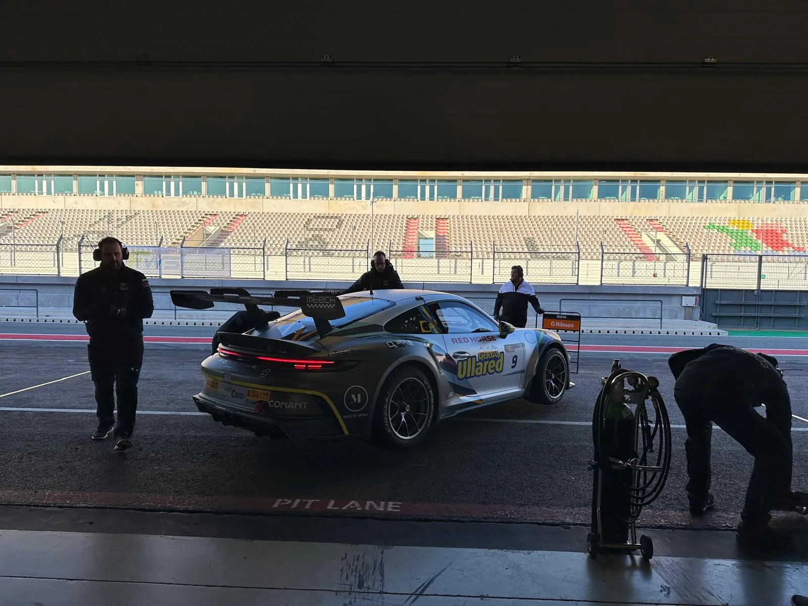The car in the pit lane at dusk with crew around it, empty grandstands behind